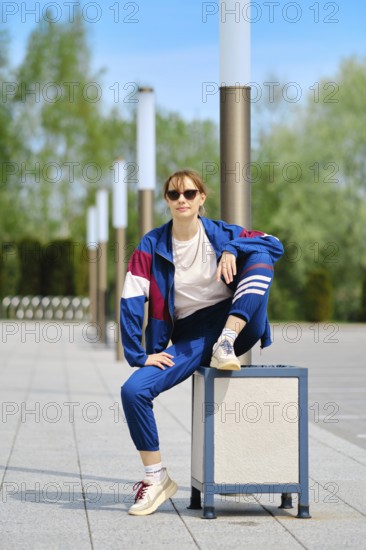 A woman dressed in a blue and maroon sporty outfit sits on a trashcan with one leg raised,