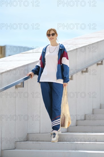A young woman with sunglasses descends a staircase wearing a colorful sporty outfit. She carries a