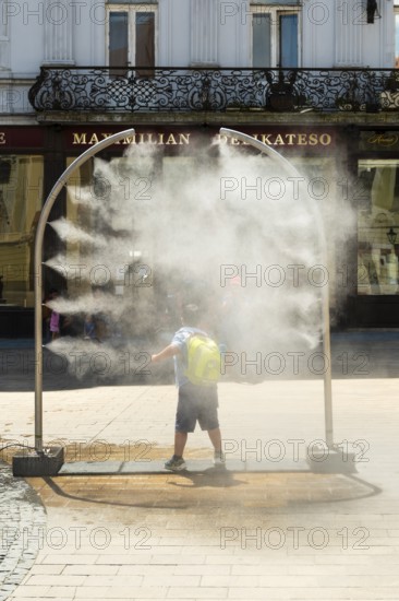 A mobile spray arch sprays a fine mist of water in a sunny square. A child uses the refreshment in