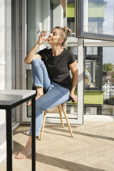 A middle-aged woman sits comfortably on a wooden chair in a sunny balcony, sipping from a glass.