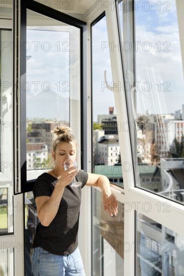 A woman leans against a balcony railing, sipping a drink and looking out at the vibrant cityscape.