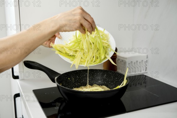 A hand pours freshly cut zucchini noodles into a sizzling pan on an electric stovetop. The kitchen