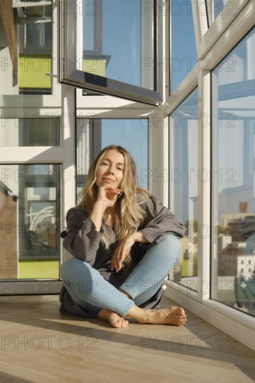 A middle-aged woman sits comfortably on a wooden floor of balcony in a bright modern apartment. She
