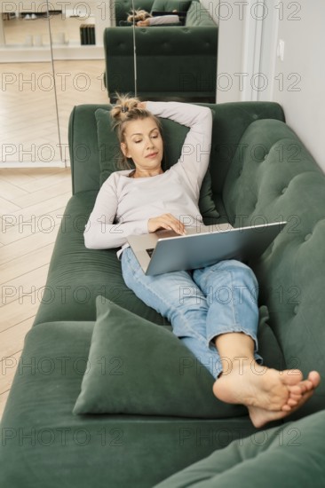 Middle-aged woman is lounging on a green couch, scrolling on her laptop in online shop choosing