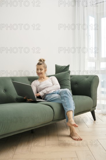 A woman enjoys her time on a green couch, focused on her laptop. Sunlight fills the bright living
