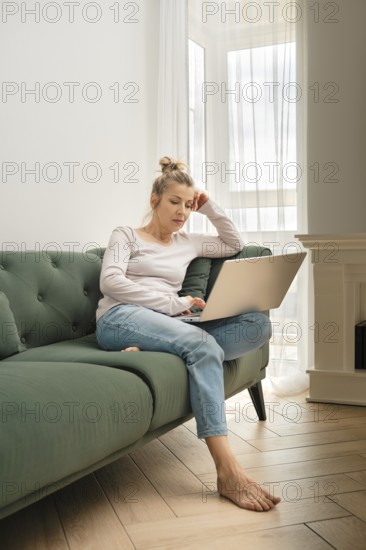 A woman is focused on her laptop while seated on a soft green sofa. She appears thoughtful, with