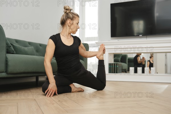A middle-aged woman in athletic wear performs leg stretching on the floor at home, combining
