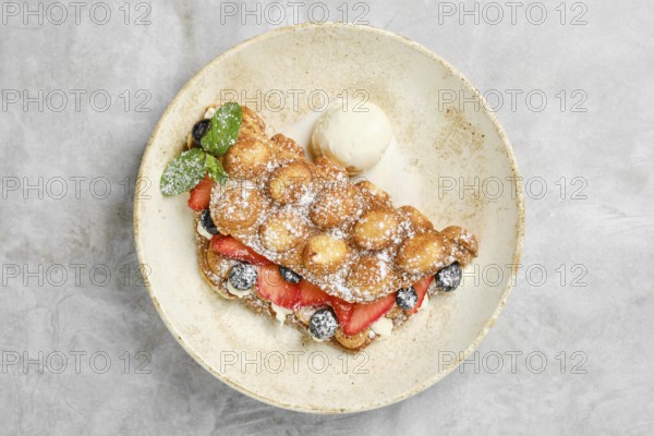 A golden bubble waffle is plated with strawberries, blueberries, and a scoop of vanilla ice cream.