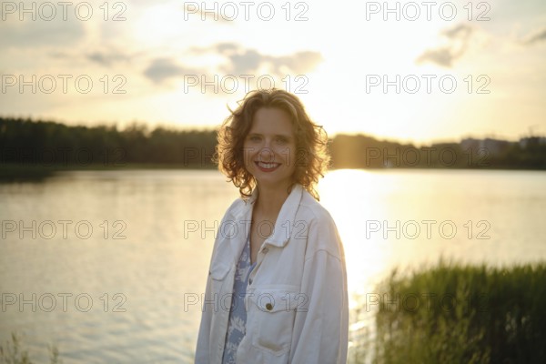 A woman stands near a shimmering lake on a hot evening, her curly hair catching the sunlight. The