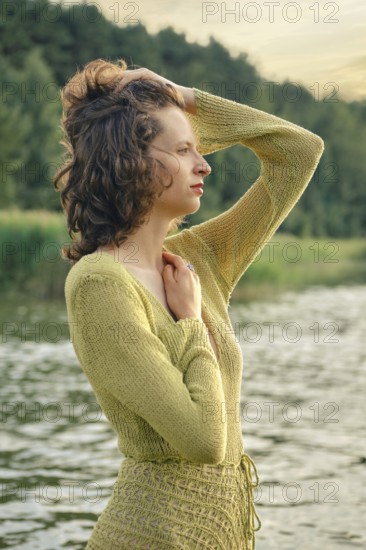 On a hot day, a woman stands gracefully by a serene lake touching her curly hair. She seems lost in