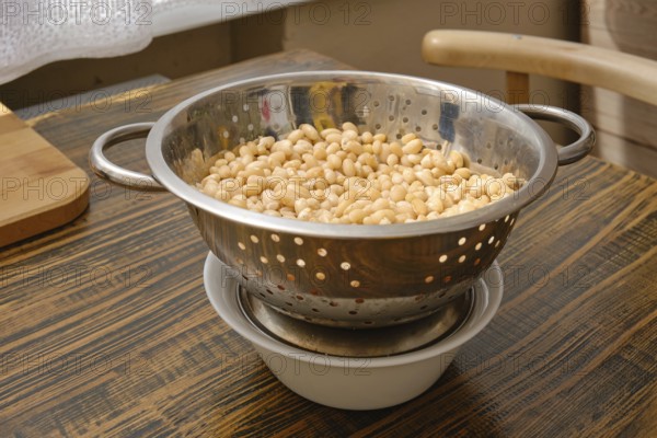 A colander filled with soaked white beans is resting above a bowl to catch excess water. Natural