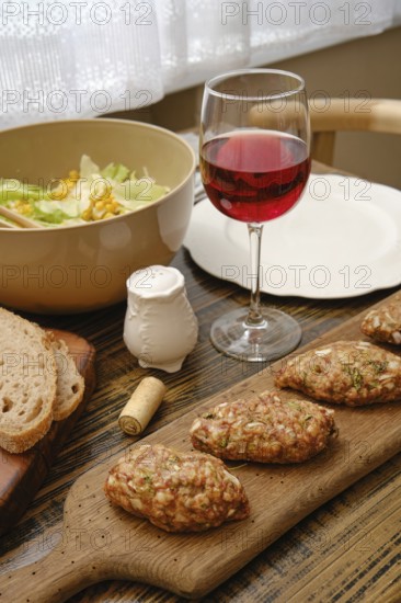 A table is set with a wooden board of homemade patties, a bowl of fresh salad, a glass of red wine,