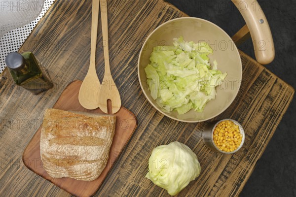A rustic wooden table displays a loaf of bread, a bowl of chopped lettuce, and a cup of corn.