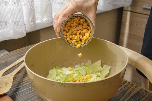 A hand pours canned corn into a bowl filled with fresh green lettuce. The warm kitchen features
