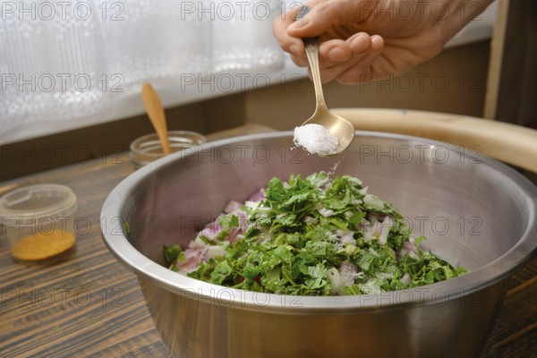 A hand adds salt to a bowl filled with minced meat, chopped onions and fresh herbs. The kitchen is