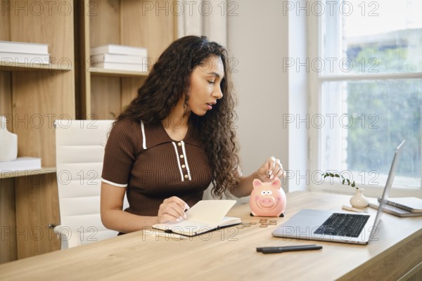 A woman sits at a wooden desk in a stylish home office, writing in a notebook while dropping coins