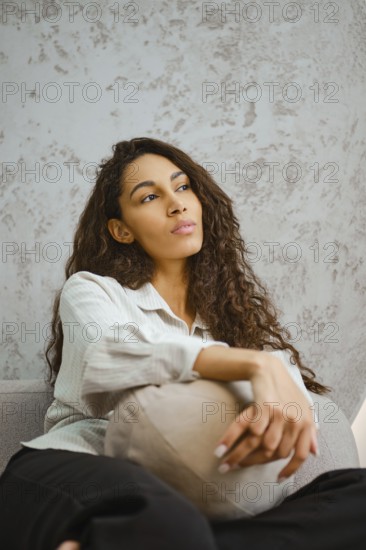 Dreamy woman sits comfortably on a gray sofa, holding a decorative pillow. Her long curly hair