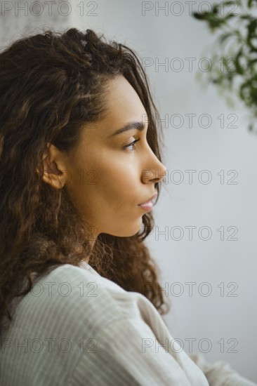 In a cozy indoor space, a woman with long, curly hair sits in profile, lost in thought. Soft