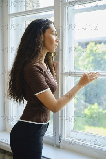 A woman admires the greenery outside while standing by a window. Her curly hair cascades down her