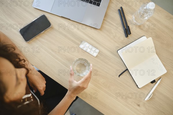 A person sits at a desk with a laptop, holding a glass of water in one hand. A pack of pills and a