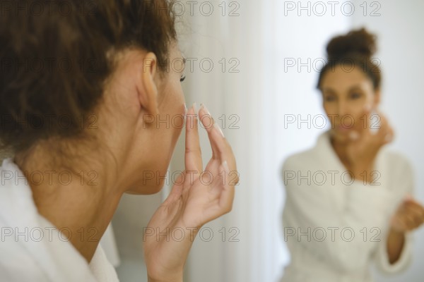 A woman in a white robe is applying cream to her face while looking at herself in the bathroom