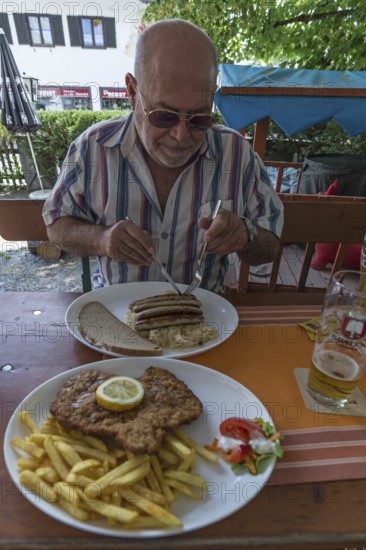 Senior citizen with sunglasses eating bratwurst in a beer garden, in front a Wiener Schnitzel with