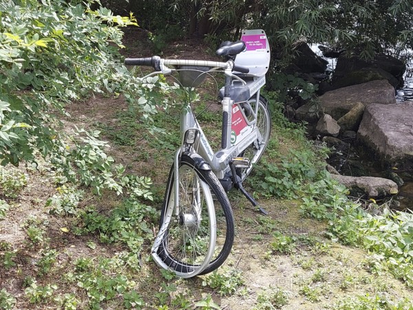 A vandalised bicycle, e-bike from a bike rental company stands in the countryside next to a river,