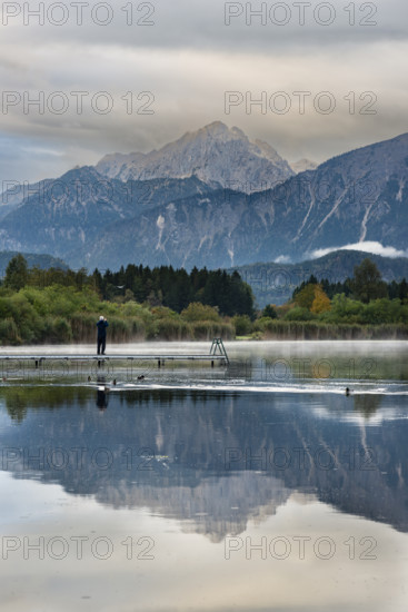 Man (60-70) with dog on a wooden footbridge at sunrise, Hopfensee, near Füssen, Ostallgäu, behind