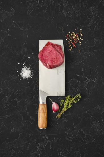Fresh meat sits atop a large knife on a dark countertop, surrounded by salt, pepper, herbs, and