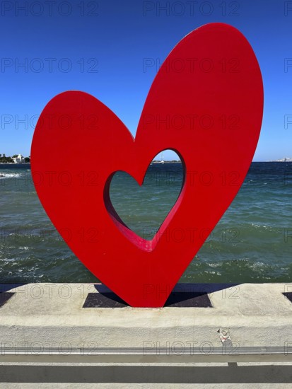 Red heart sculpture in front of blue sea and sky, romantic modern art on concrete base, Kos Town,