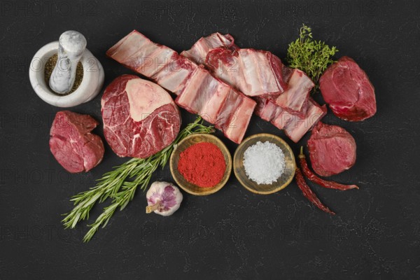 A variety of fresh cuts of beef are arranged on a dark surface, surrounded by herbs, spices, and
