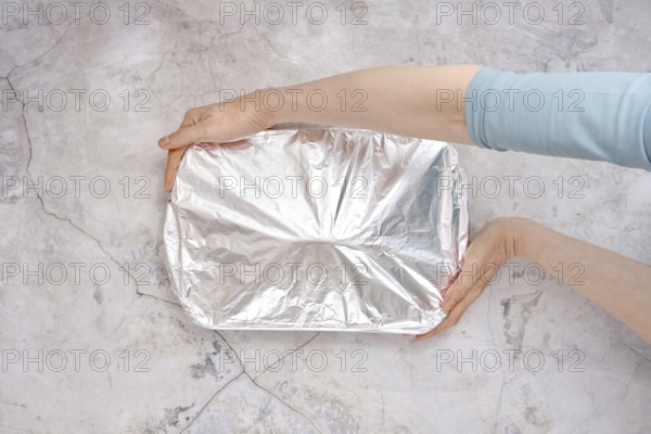 A hand holds a baking dish covered with aluminum foil, ready for the oven. The setting features a