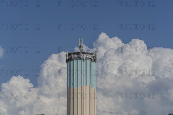 A tall water tower, painted in bright colours, rises against a backdrop of flowing white clouds.