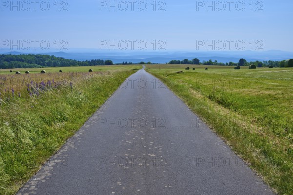 Straight road through a vast landscape of green meadows and distant hills under a blue sky,