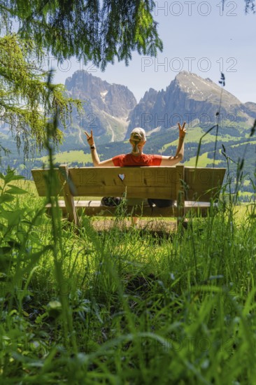 Person on a bench showing a peace sign in front of a mountain landscape, Alpe di Siusi, Dolomites,