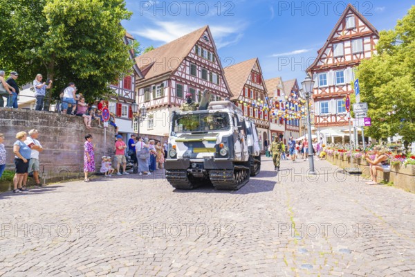 A military vehicle drives on a festive street with half-timbered buildings and people passing by,