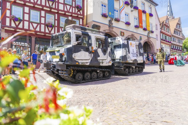 Two military vehicles in front of a row of historic half-timbered houses and surrounded by blooming