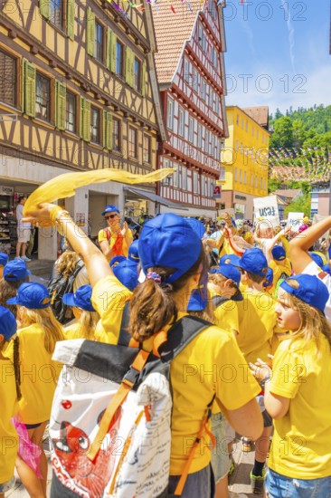 A group of children in yellow shirts and blue caps celebrates in an old town with half-timbered