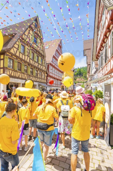 A colourful parade with yellow balloons moves through lively streets lined with half-timbered