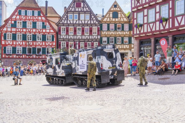 Armoured vehicles stand on a town square with half-timbered buildings and soldiers in camouflage,