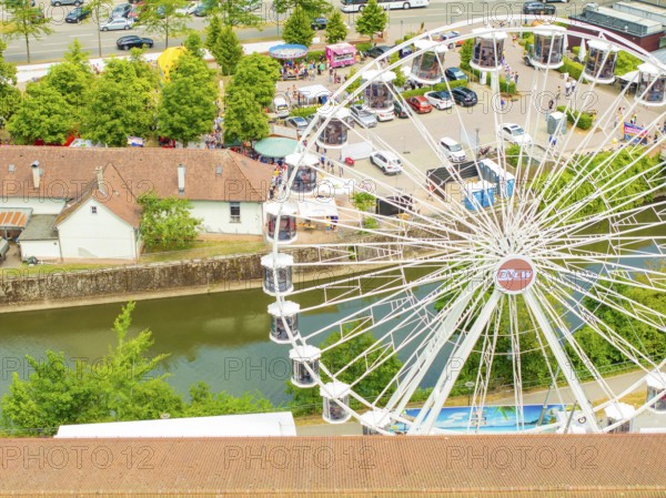 Ferris wheel stands next to a river and car parks, surrounded by trees in summer, 950 years of