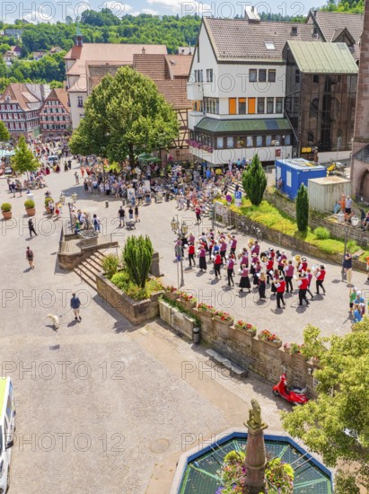 A parade moves through a market square, surrounded by half-timbered houses and many spectators in
