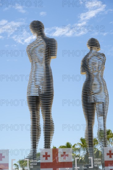 Metal figures of a woman and a man against a blue sky, Georgian flags at the bottom of the picture,