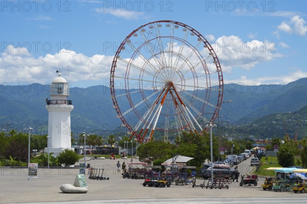 Big Ferris wheel and white lighthouse with mountains in the background under blue sky, amusement