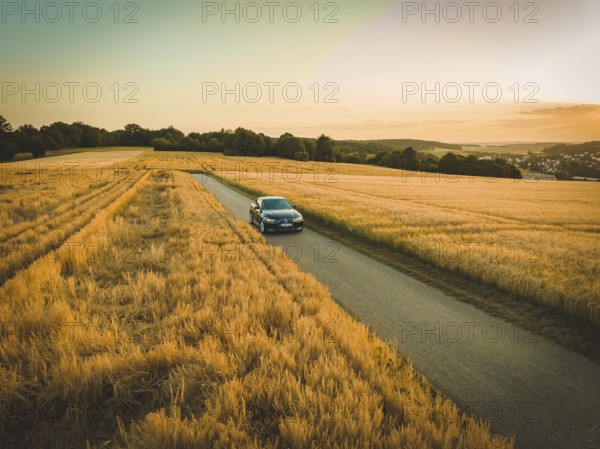A car drives on a country road between golden fields at sunset, BMW i7, Deer E-Carsharing, Calw,