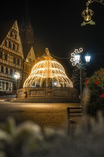 Illuminated fountain in front of half-timbered houses surrounded by festive Christmas lights at