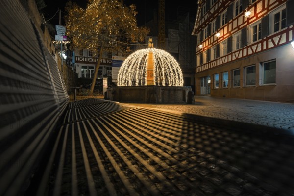 Night view of a festively illuminated fountain with shadows cast in a historic street, Calw, Black