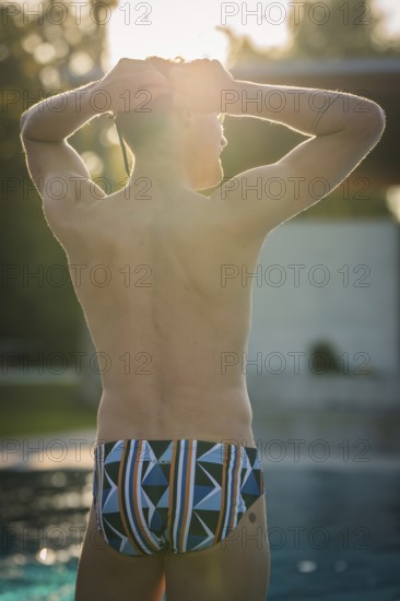 A man in swimming trunks stands by the pool in sunlight and enjoys the relaxed atmosphere,