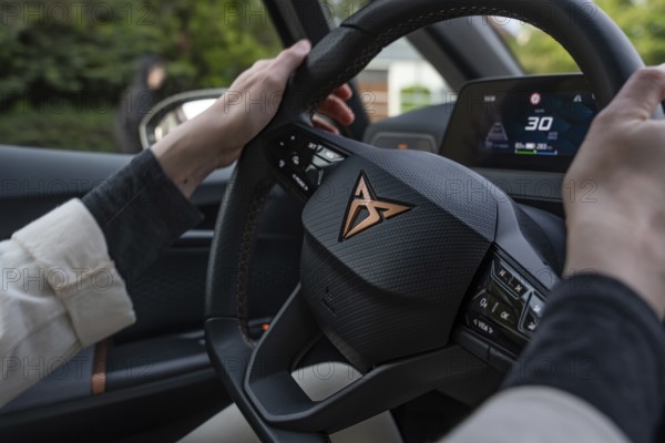 A person holds a Kupra steering wheel while the driver sits in the car and shows the dashboard with