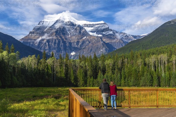 Two tourists standing on a wooden deck are enjoying the breathtaking view of mount robson, the